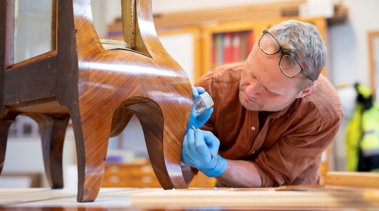 På ett bord står ett träföremål. En person står lutad över det och kikar på en detalj med lupp.