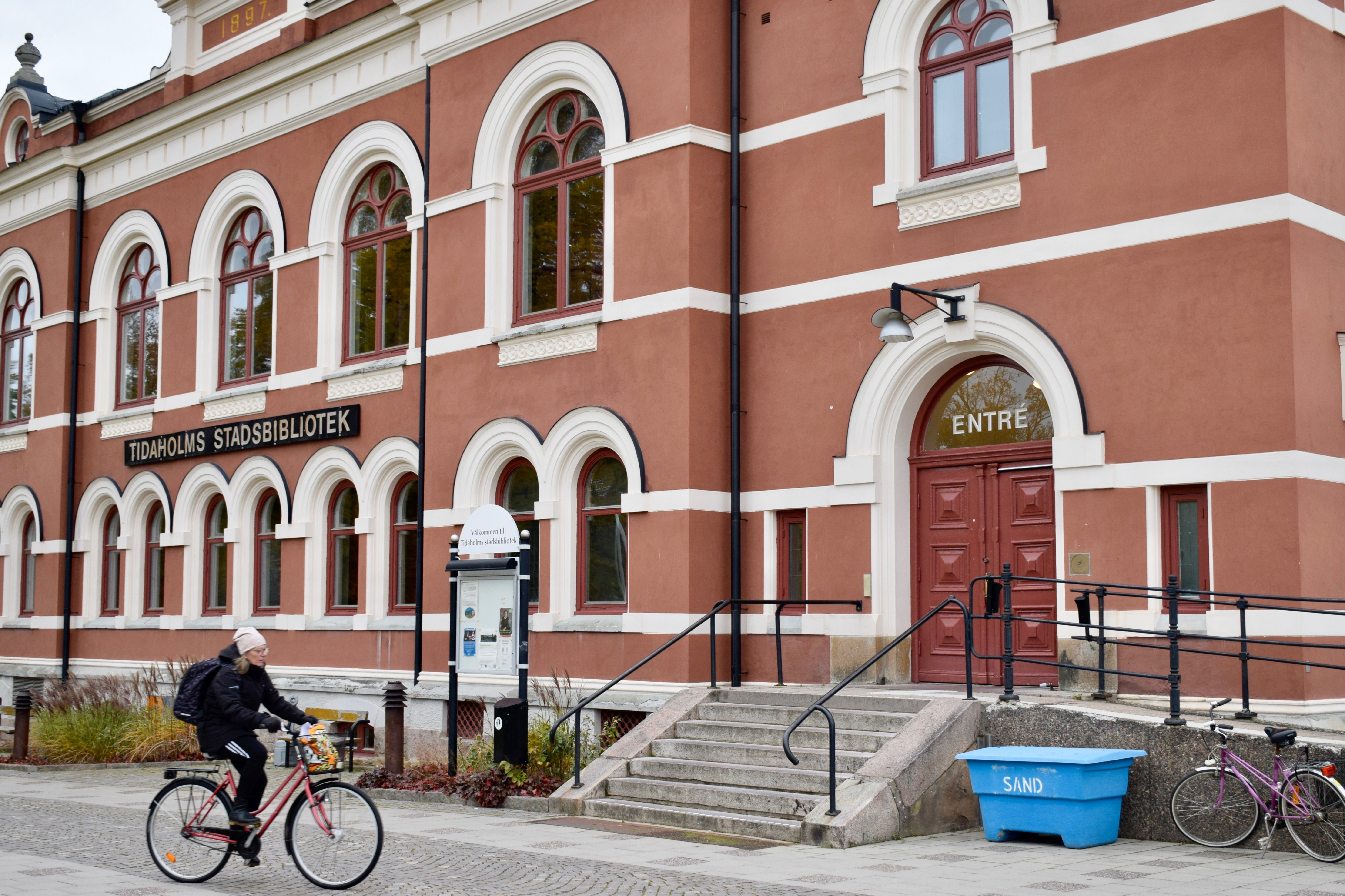 Tidaholms bibliotek, en röd byggnad med vita fönster som är formade som bågar längst upp. En kvinna cyklar förbi, på huset står skylten TIDAHOLMS BIBLIOTEK.