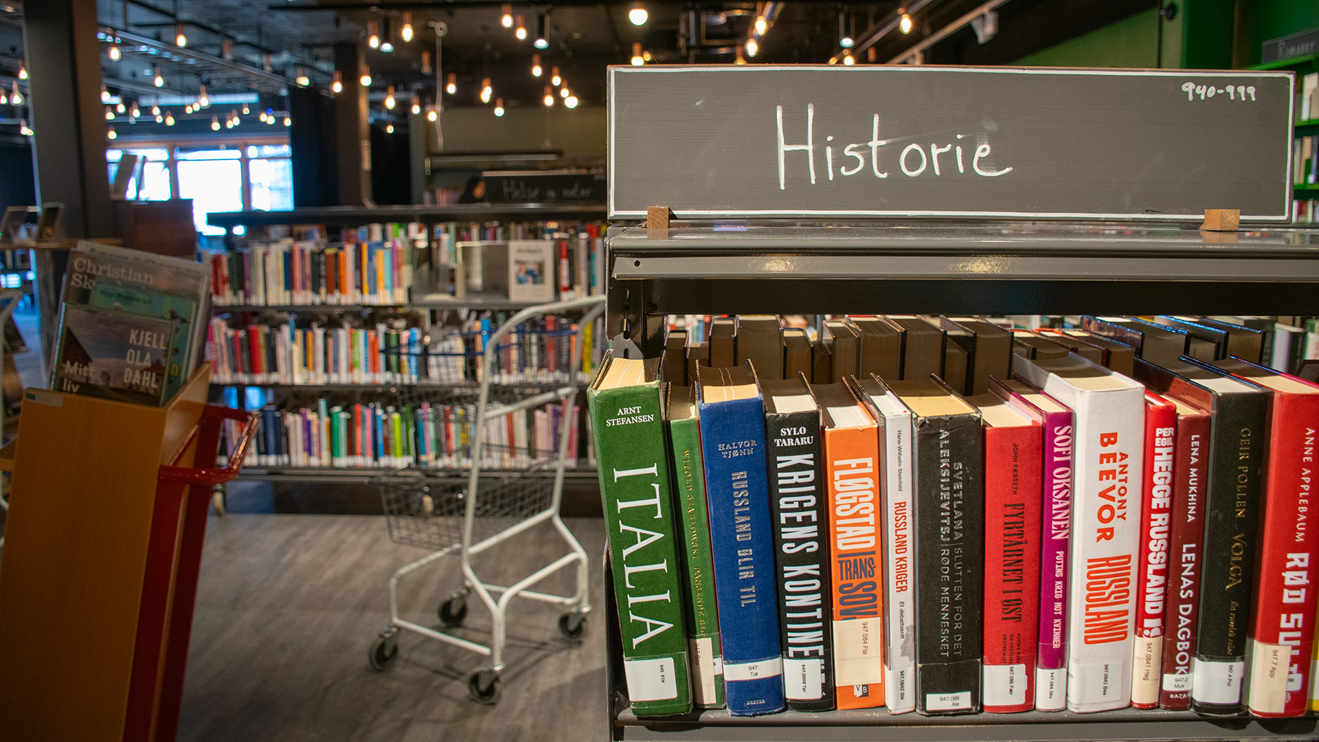 En bokhylla fylld med böcker med skylten "Historie". Bredvid hyllan står en mobil bokvagn och en vagn med två metallkorgar som är tom. 