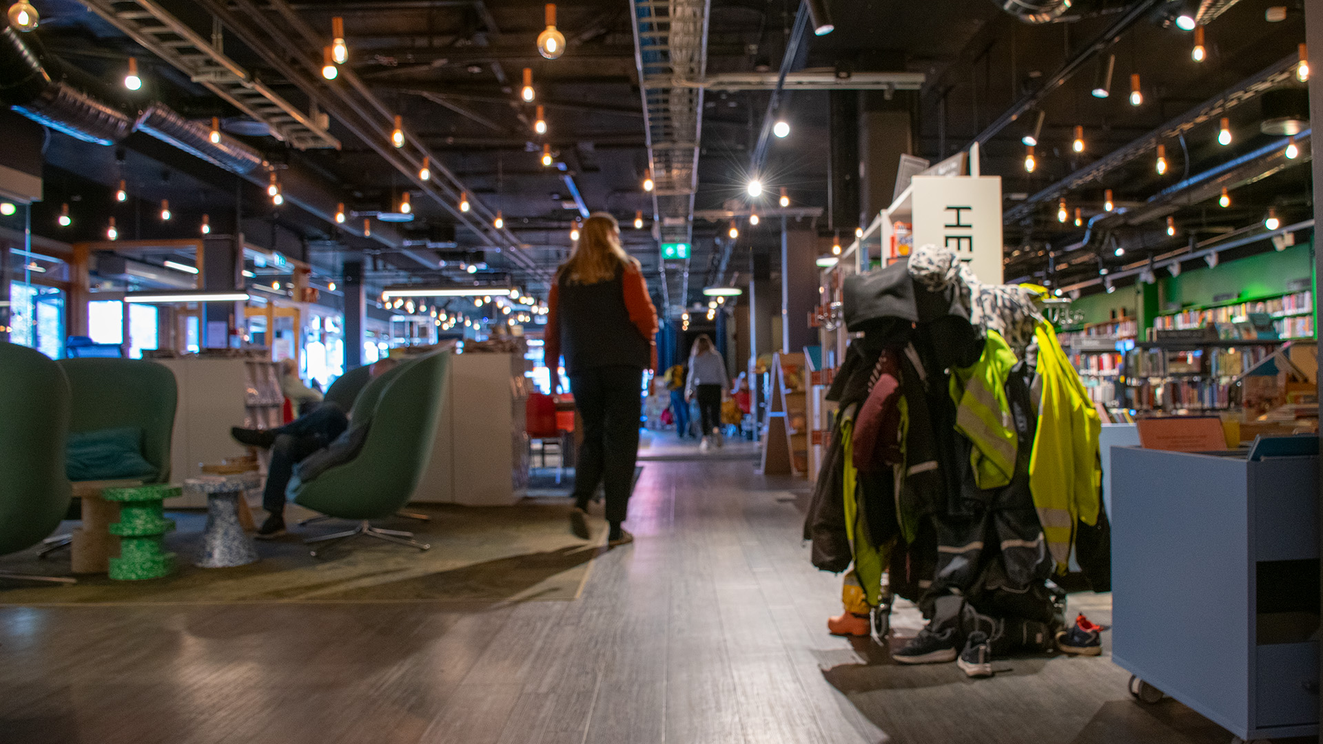 En rak gång i biblioteksrummet. Till vänster finns ett antal fåtöljer med hög rygg, någon sitter i en av dem. En hel hög med kläder och varselvästar från besökande förskolebarn hänger till höger. I taket hänger stativ och på dem små spotlights. 