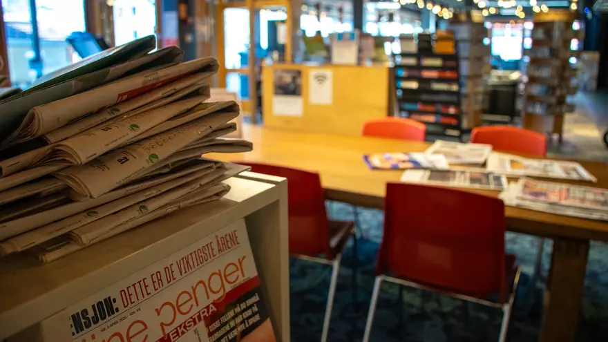 En hög med dagstidningar ligger på en hylla i förgrunden. Ett stort bord med röda stolar finns längre bort i rummet., på bordet ligger flera dagstidningar utspridda.