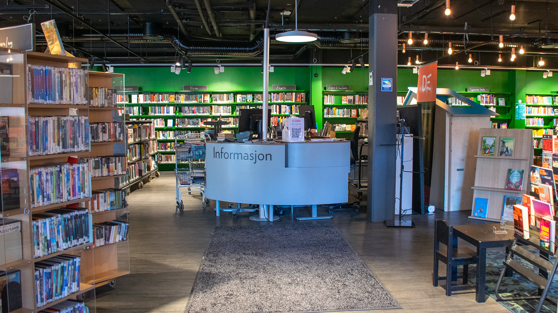 I mitten av bilden finns bibliotekets informationsdesk. Den är vit och har stor text: "Informasjon". En matta och en rak gång leder fram till desken. Runtom finns portabla hyllor med hjul, väggen i bakgrunden är målad i en klar grön färg.