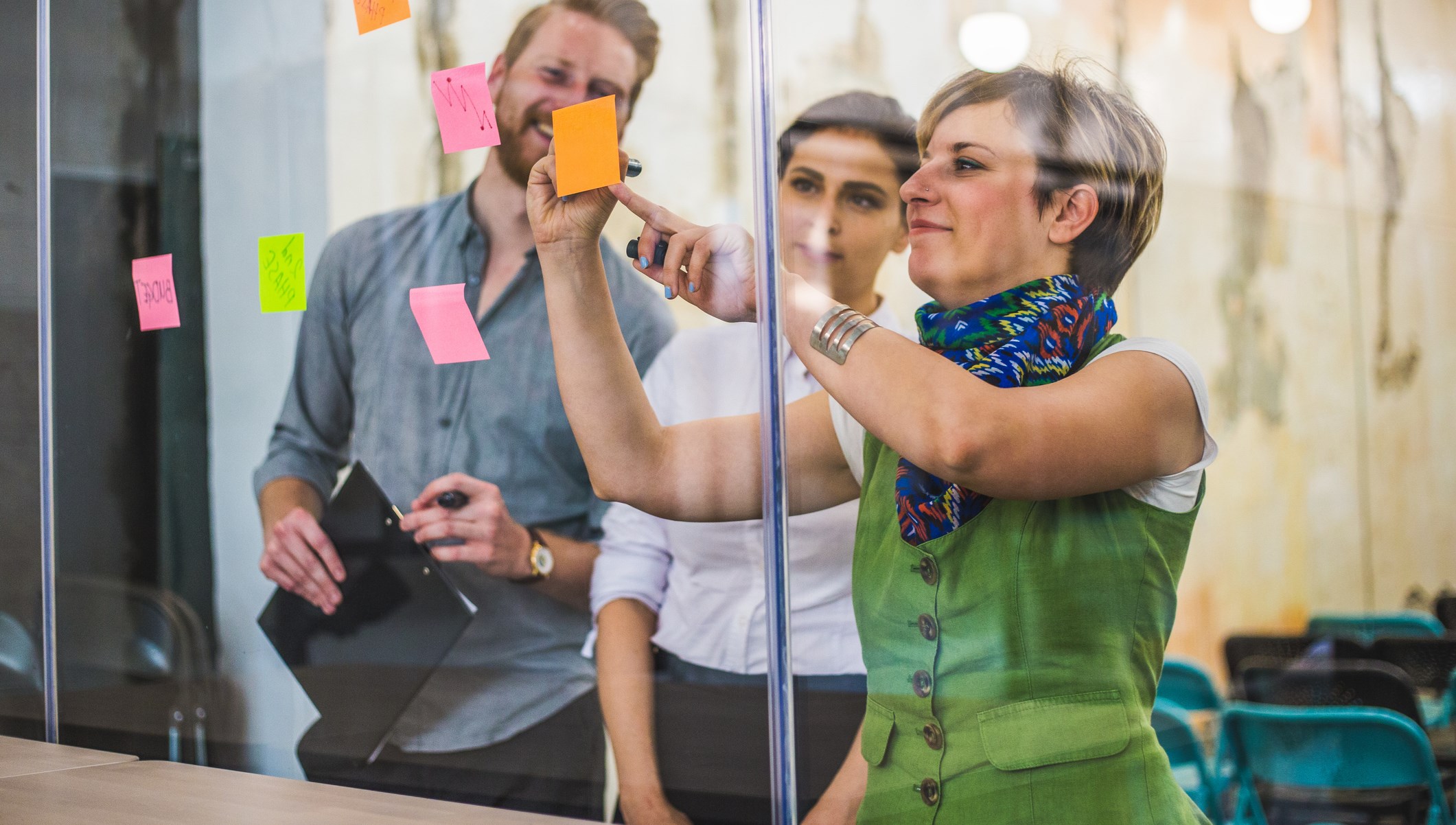 Två kvinnor och en man sätter upp post-itlappar på en glasvägg på arbetsplatsen