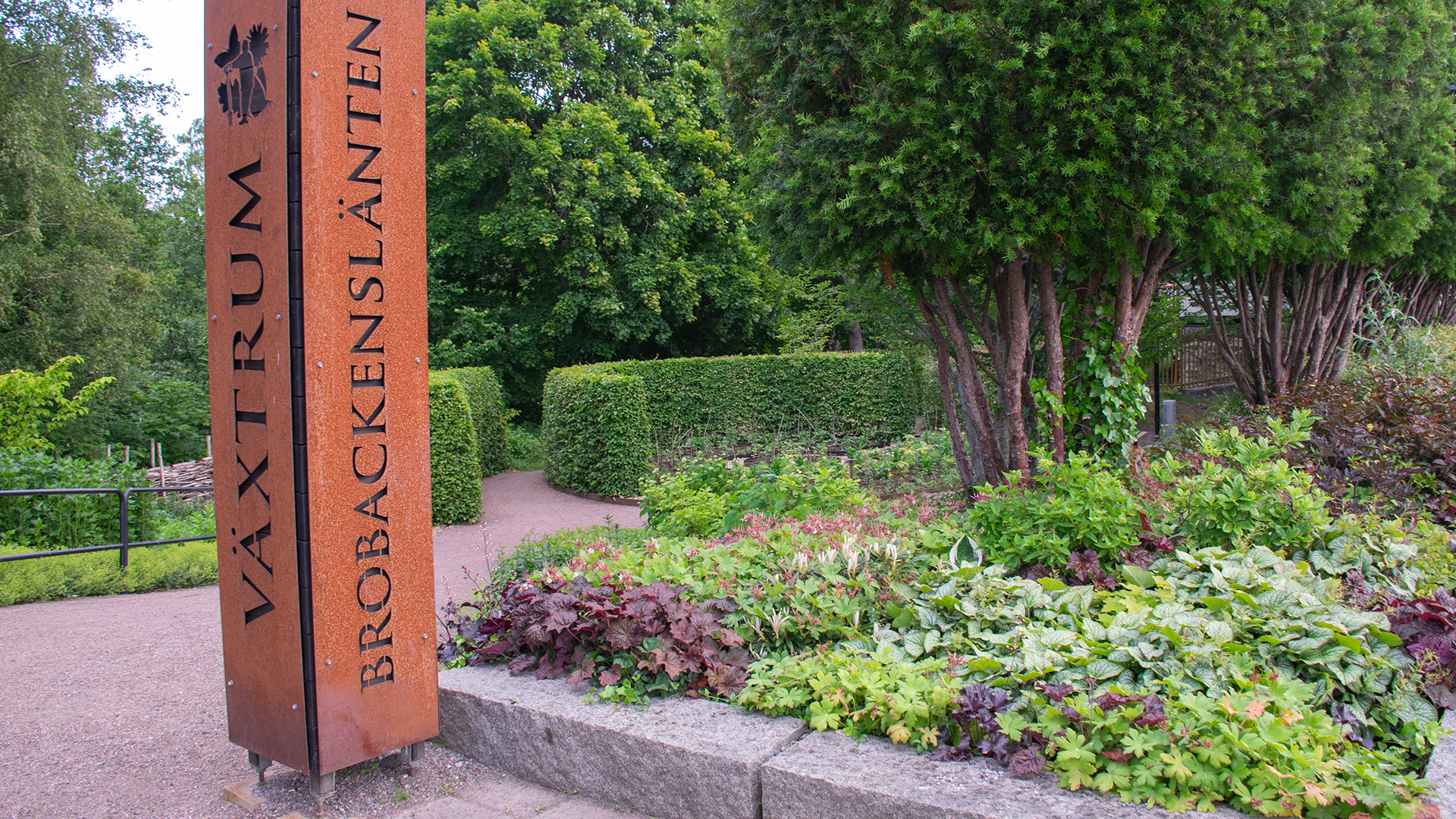 En rabatt med växter, träd i rad och en formklippt häck. En stor stiliserad skylt i rödrostig metall markerar ingången till den lilla parken. På skylten står det "Växtrum Brobackenslänten". 
