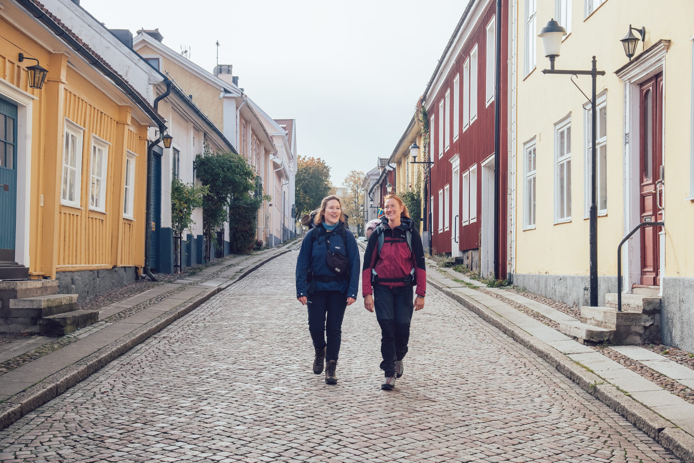 Två kvinnor passerar färgglada trähus på en kullerstensgata.