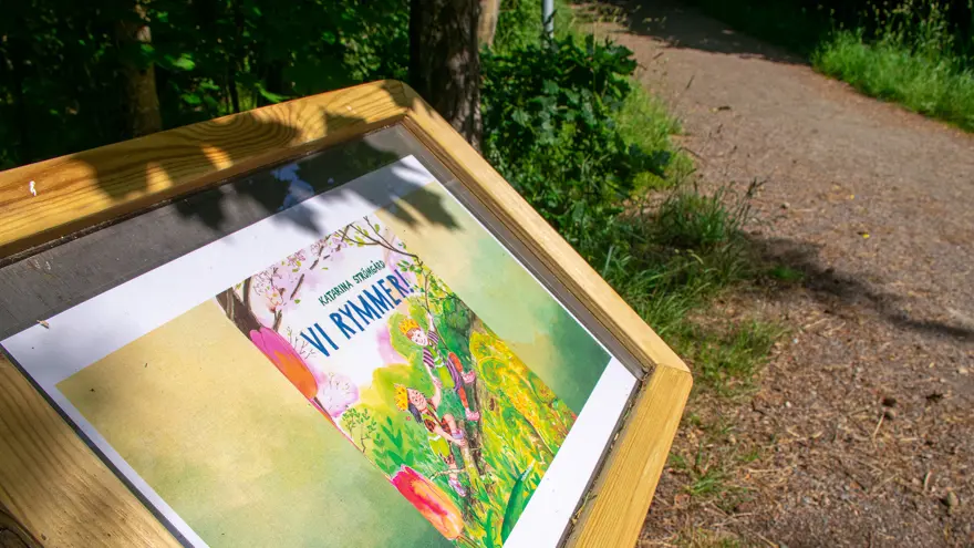 En låg skylt med träram vid sidan av en gångväg i skogen. Innanför glaset finns framsidan till boken Vi rymmer.