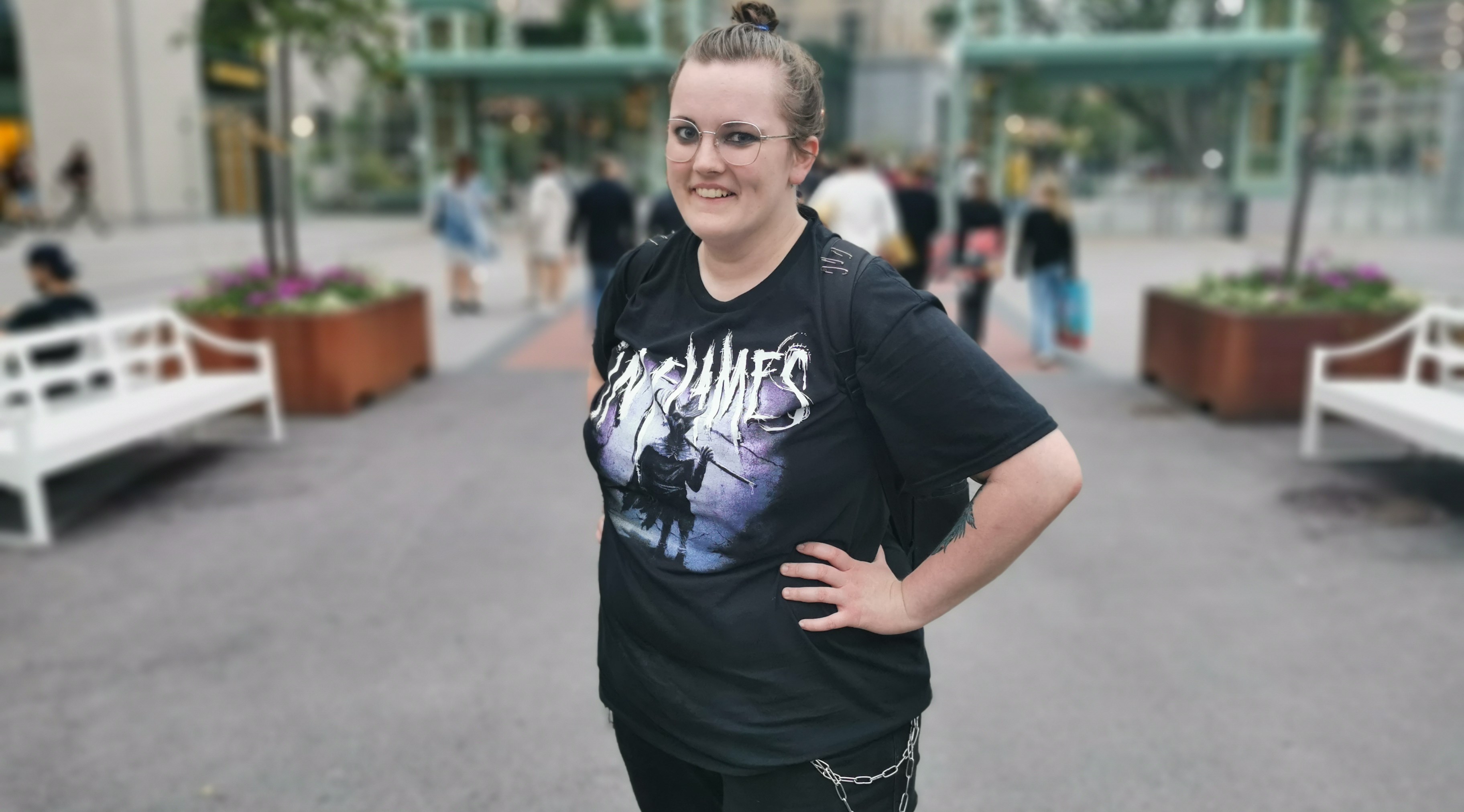 Kvinna i svarta kläder. InFlames tryck på t-shirten. Tofs mitt på huvuudet. Glasögon med tunna bågar. Metallkedja hänger i de svarta jeansen. 