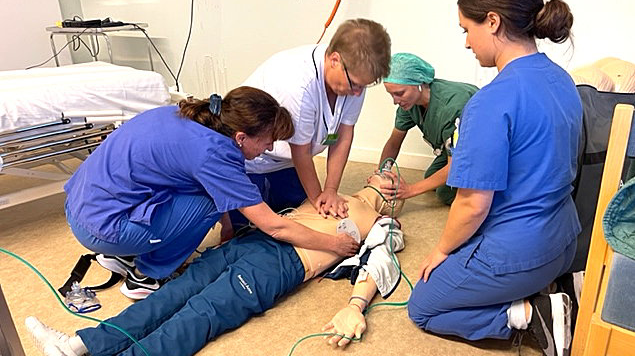 Tre medarbetare övar HLR på en docka. En instruktör observerar.