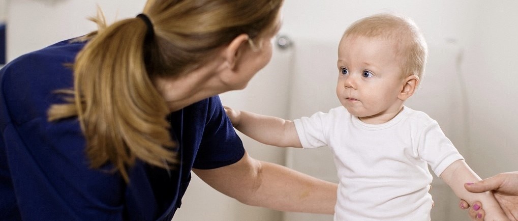  En kvinna i blå läkarrock tittar på ett litet barn som bär en vit body. Vi ser kvinnans rygg och hästsvans och barnet framifrån. Barnet sitter själv men kvinnan stöttar med sin arm bakom barnets rygg. 