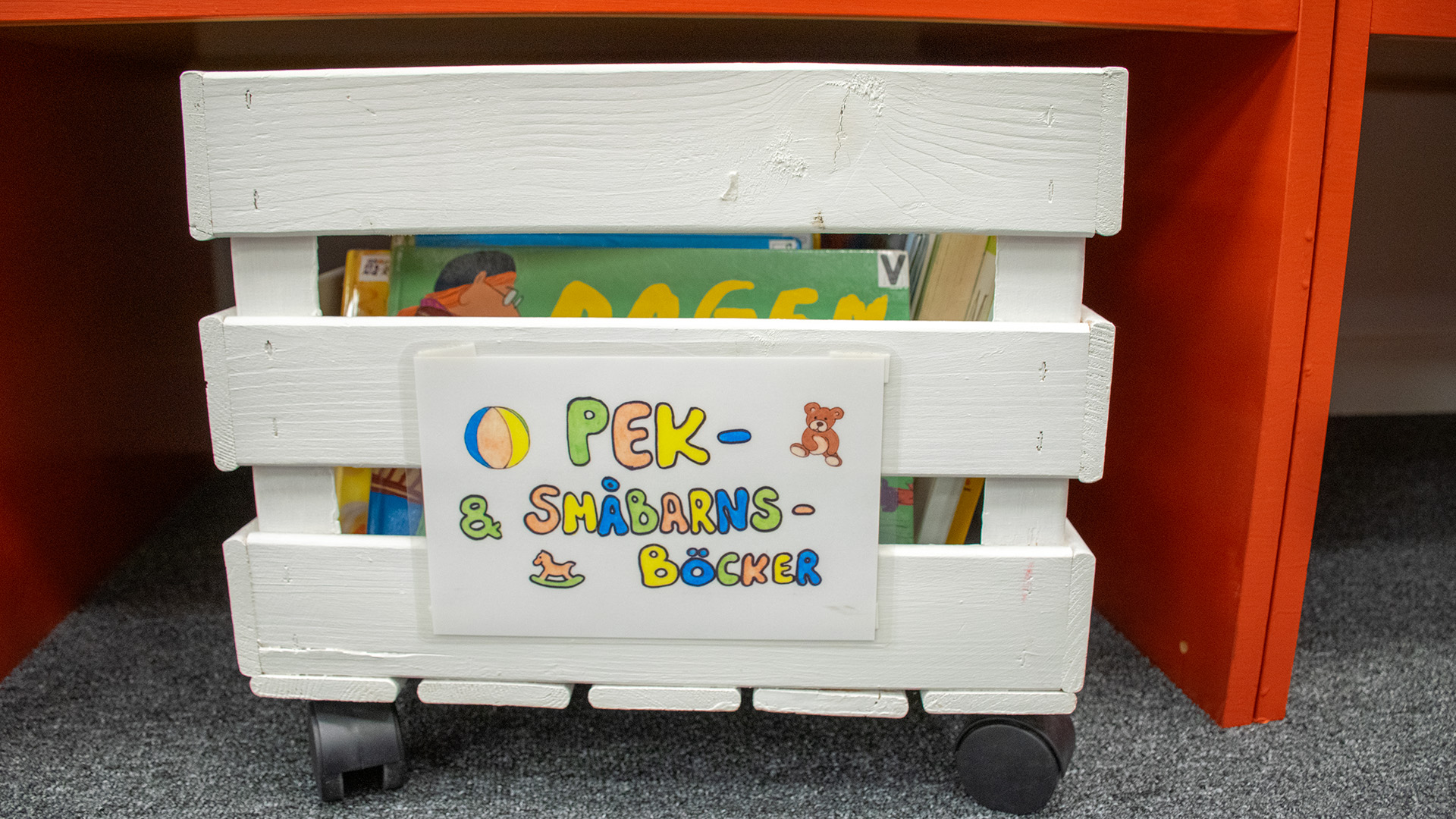 En vitmålad träback på hjul, fylld med böcker. Med färgglada bokstäver står det på en skylt Pek- & småbarnsböcker.  