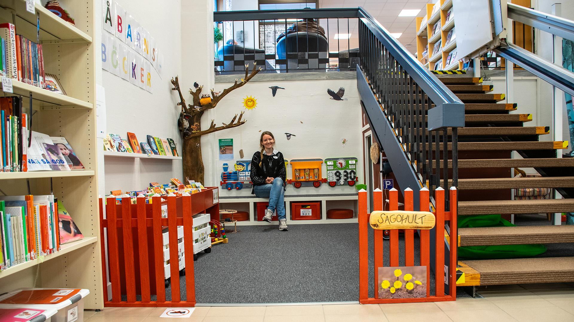 Småbarnsavdelningen Sagohult. Avdelningen är en hörna till vänster om en trappa. Vita väggar, ett träd, boktråg och en grå heltäckningsmatta på golvet. 