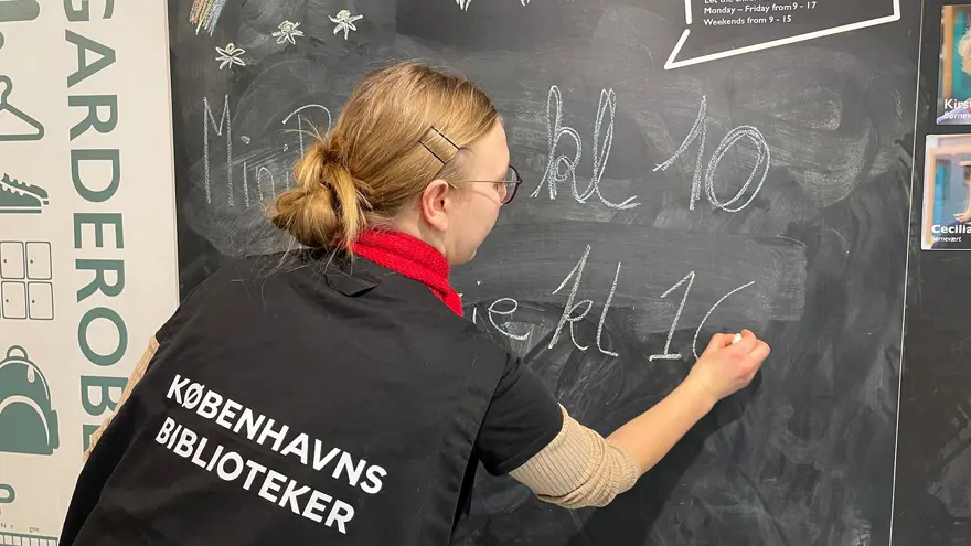 En bibliotekarie skriver dagens program på en svart tavla. Bibliotekarien har en svart väst med texten Köpenhamns bibliotek på ryggen.